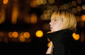 A woman with short blonde hair wearing a black coat, shown in profile against a dark background with warm yellow bokeh lights.